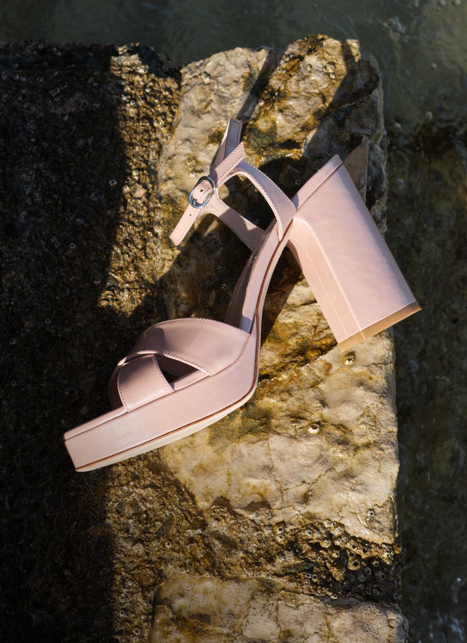 Dayna Platform Sandal 110 in blush pink leather with a block heel, placed on coastal rocks beside the water. 