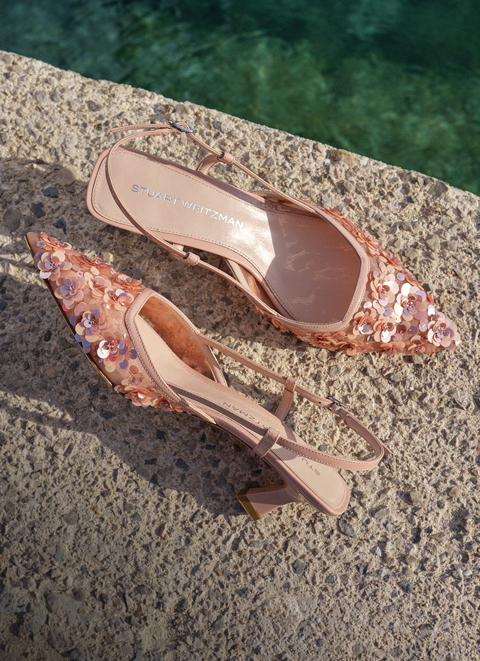 The Vinnie Slingback Pump in blush pink with floral sequin embellishment, styled on a stone surface beside a pool. 